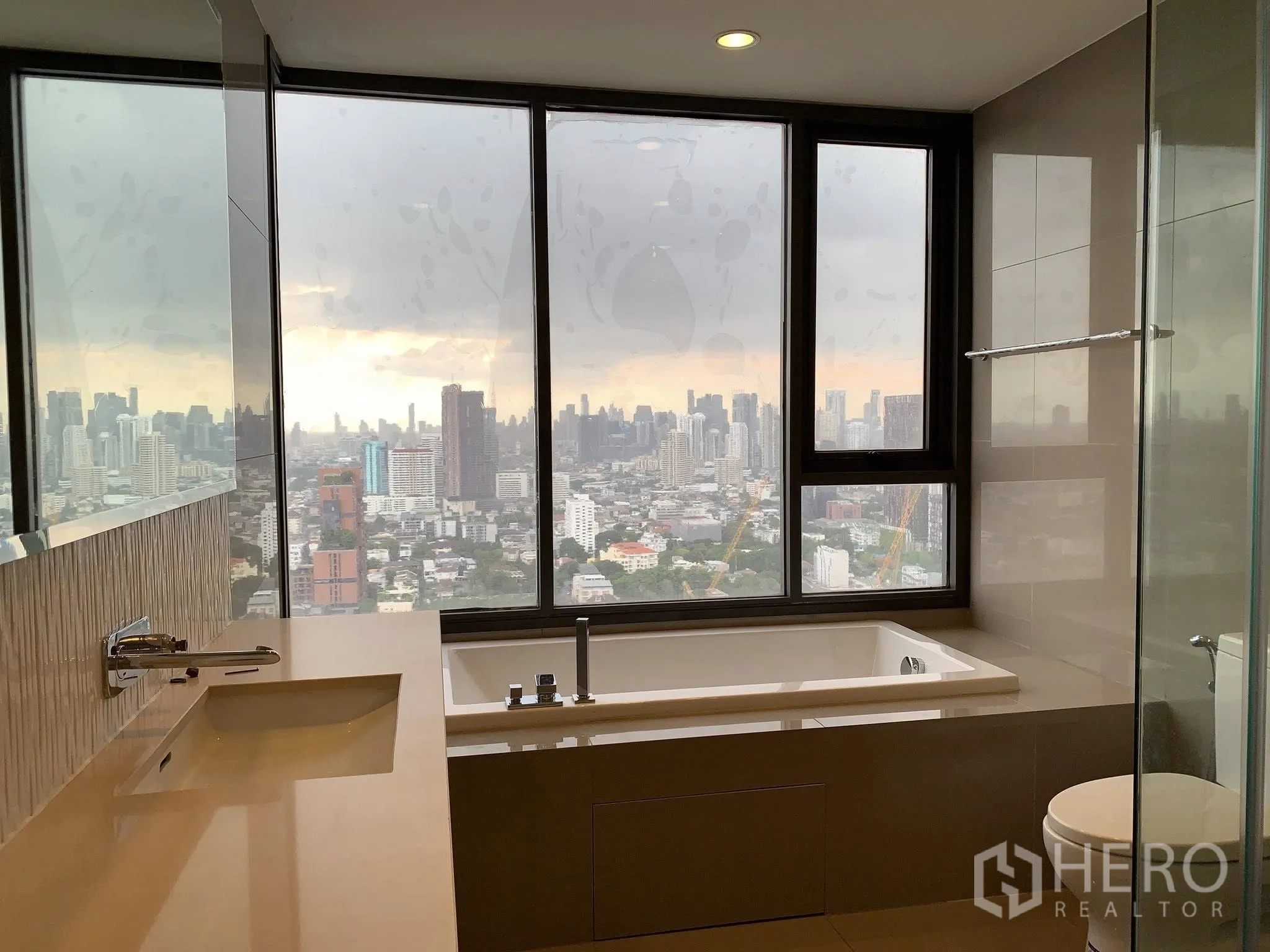 Condominium for rent in Khlong Toei, Bangkok - Bathroom with large window, built-in bathtub, vanity, and expansive city skyline view.