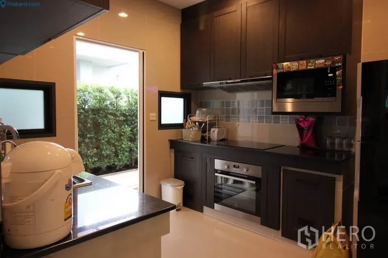 Detached House for rent or sale in Prawet, Bangkok - Fitted kitchen with dark cabinetry, built-in oven and microwave, opening to the side garden.