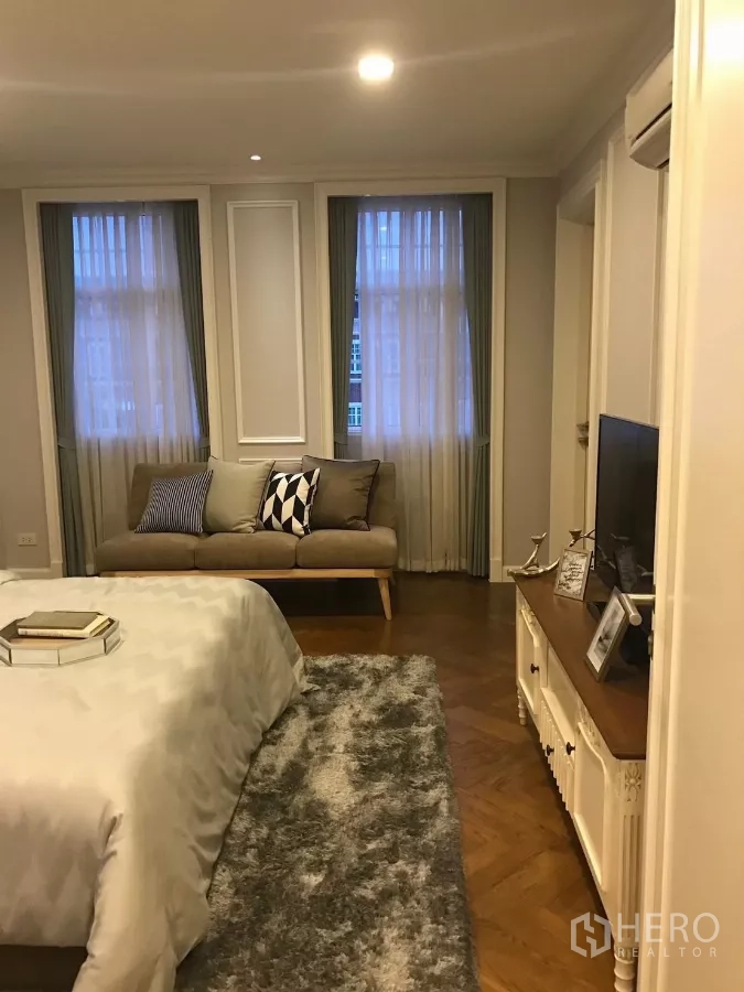 Townhouse for sale in Watthana, Bangkok - Bedroom lounge area with sofa, TV console and tall curtained windows.