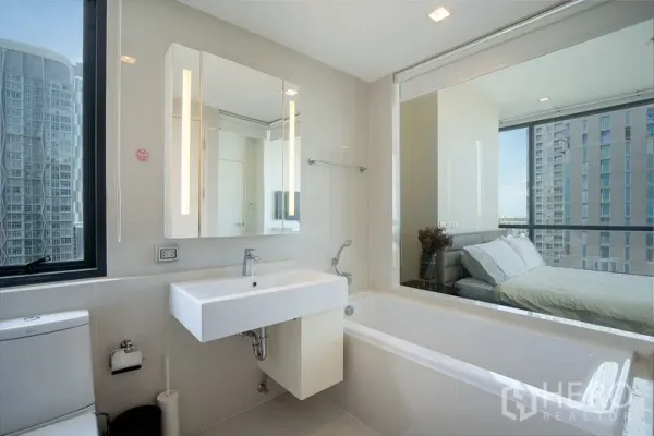 公寓 出租在Watthana, Bangkok - 明亮浴室带浴缸与洗手台，玻璃墙连接卧室，可欣赏曼谷城市景观，近On Nut。