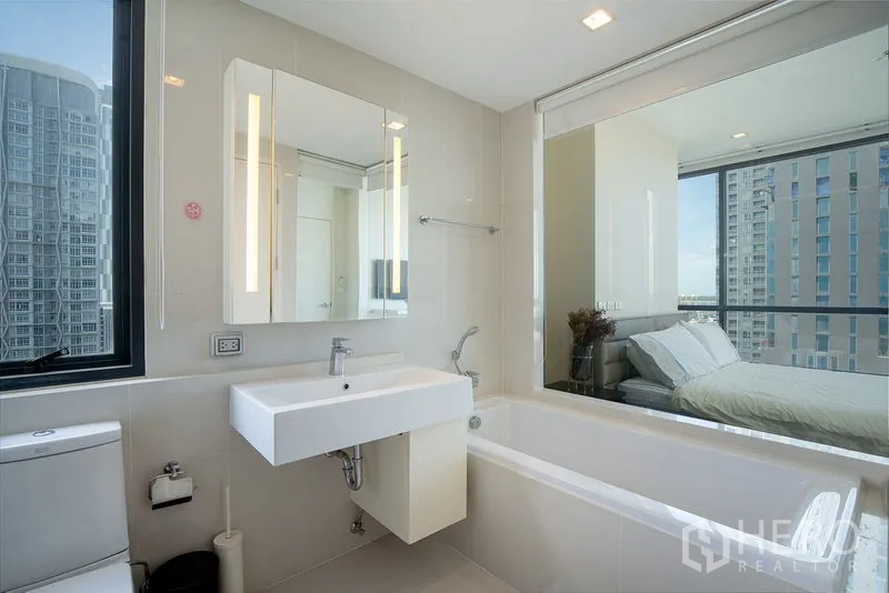 公寓 出租在Watthana, Bangkok - 明亮浴室带浴缸与洗手台，玻璃墙连接卧室，可欣赏曼谷城市景观，近On Nut。