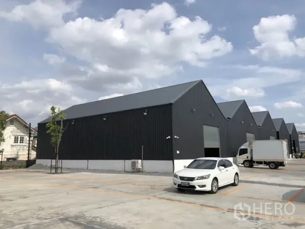 Warehouse for rent in Mueang Nonthaburi, Nonthaburi - Row of modern black metal-clad warehouses with gabled roofs, roller shutter doors, and wide concrete parking and loading area with car and truck.