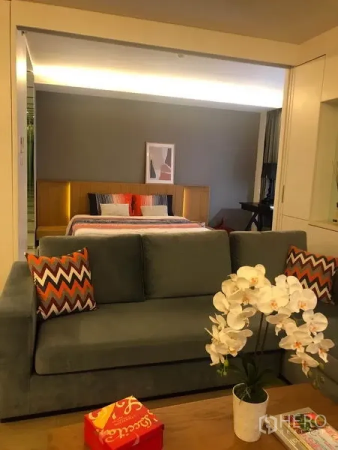 Condominium for rent in Watthana, Bangkok - Cozy living area with a gray sofa, orchid on the coffee table, and an open view to the bedroom.