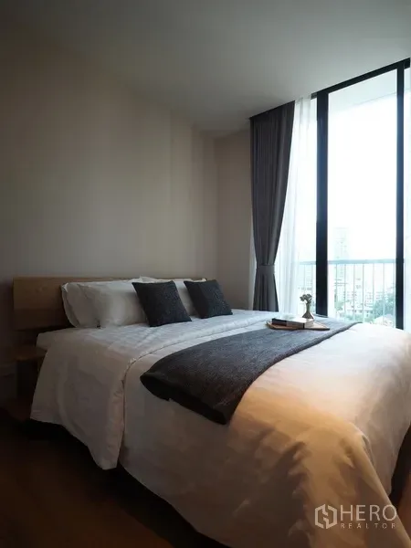 Condominium for sale in Khlong Toei, Bangkok - Cozy bedroom with a double bed, grey accents and a tall window opening to a city-view balcony.