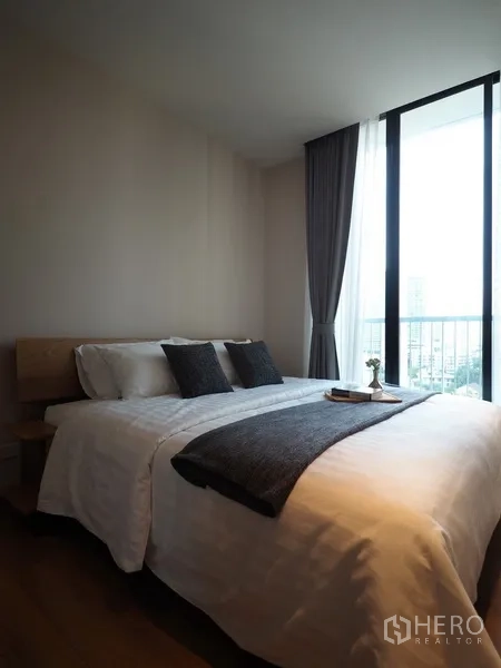 Condominium for sale in Khlong Toei, Bangkok - Cozy bedroom with a double bed, grey accents and a tall window opening to a city-view balcony.