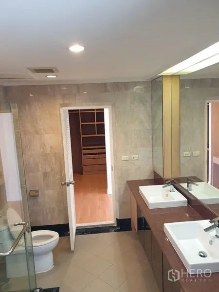 Condominium for rent in Watthana, Bangkok - Bathroom angle showing toilet, glass shower, twin basins and doorway to a walk-in closet.