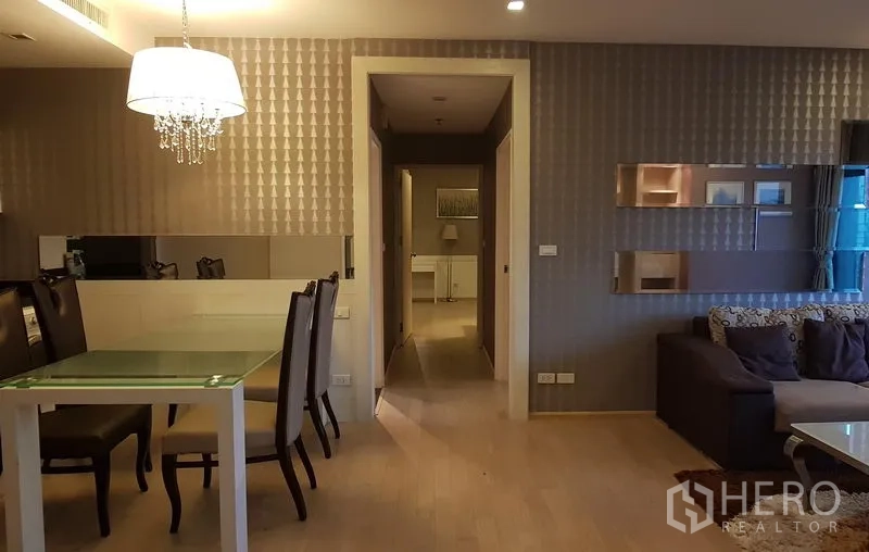 公寓 出租在Khlong Toei, Bangkok - 餐区配玻璃餐桌和吊灯，镜面装饰墙旁通往卧室走廊。