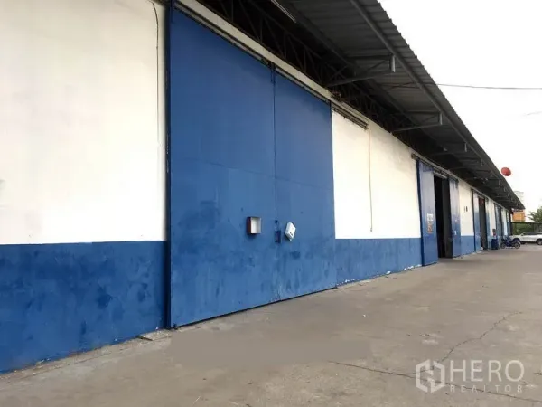 คลังสินค้า ให้เช่าในBang Phli, Samut Prakan - ด้านหน้าโกดังโทนสีน้ำเงิน-ขาว พร้อมประตูบานเลื่อนเหล็กขนาดใหญ่และหลังคาคลุมลานโหลด บนพื้นคอนกรีตกว้างในบางพลี