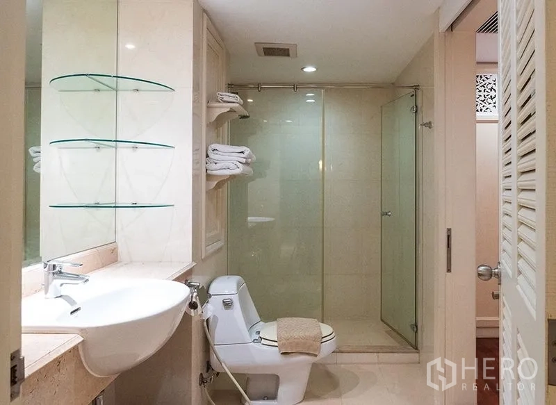 Apartment for rent in Bang Rak, Bangkok - Modern bathroom with glass-enclosed shower, vanity basin, toilet and towel shelves in neutral tones.