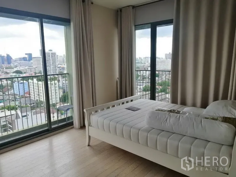 公寓 出租在Watthana, Bangkok - 卧室配白色床架，落地窗与阳台，可欣赏城市天际线景观。