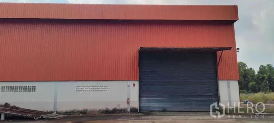 货栈 出租在Nikhom Phatthana, Rayong - 仓库正面配大卷闸门与雨棚，铺装卡车出入口。