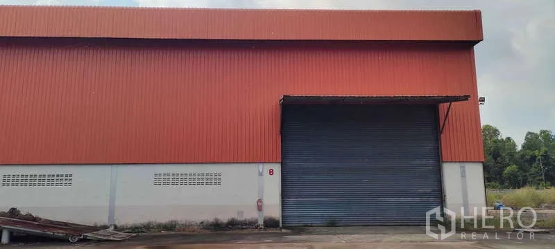 货栈 出租在Nikhom Phatthana, Rayong - 仓库正面配大卷闸门与雨棚，铺装卡车出入口。