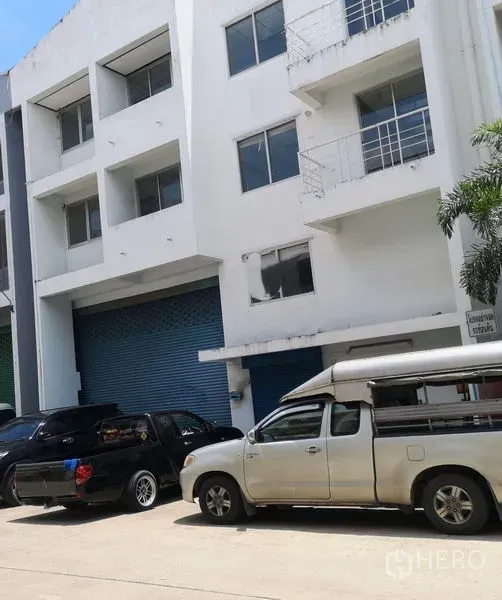 Factory for sale in Khlong Luang, Pathum Thani - Multi-storey factory facade with blue roller shutter and wide access road with parking.