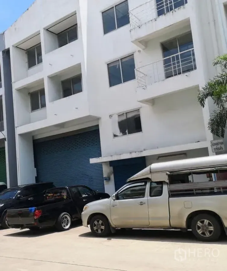 Factory for sale in Khlong Luang, Pathum Thani - Multi-storey factory facade with blue roller shutter and wide access road with parking.