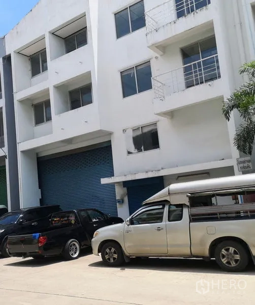 Factory for sale in Khlong Luang, Pathum Thani - Multi-storey factory facade with blue roller shutter and wide access road with parking.