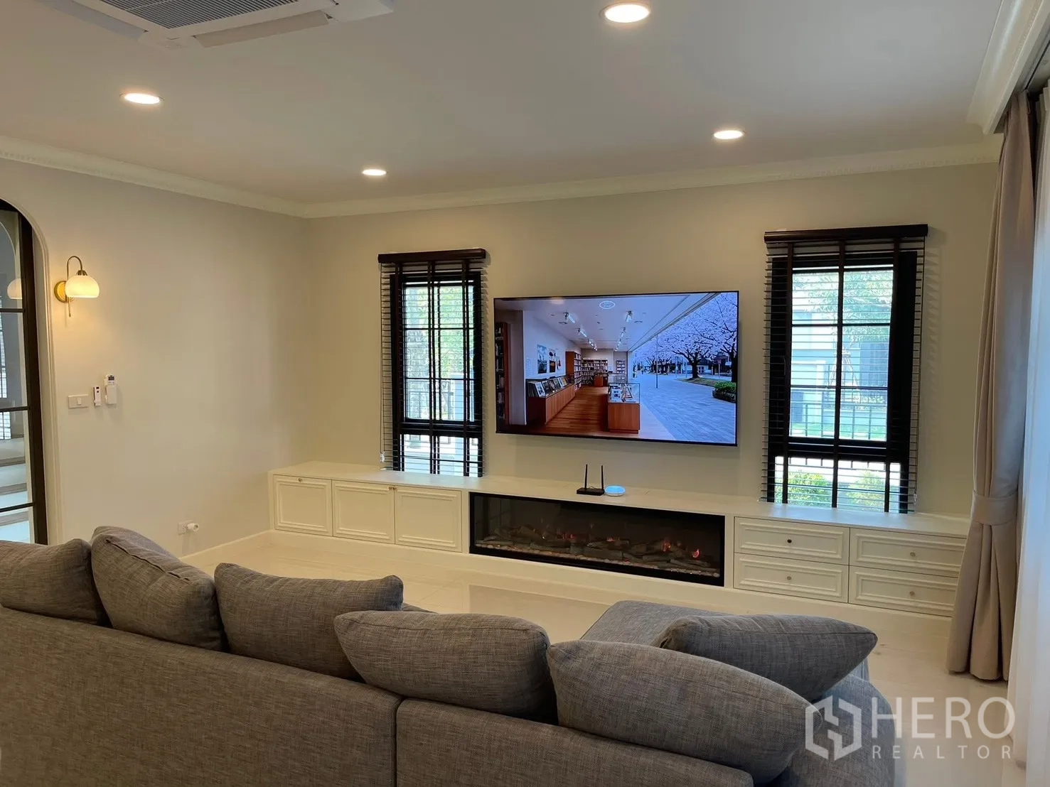 Detached House for rent in Bang Phli, Samut Prakan - Living area with large wall-mounted TV, built-in media cabinet with electric fireplace feature, and twin windows.