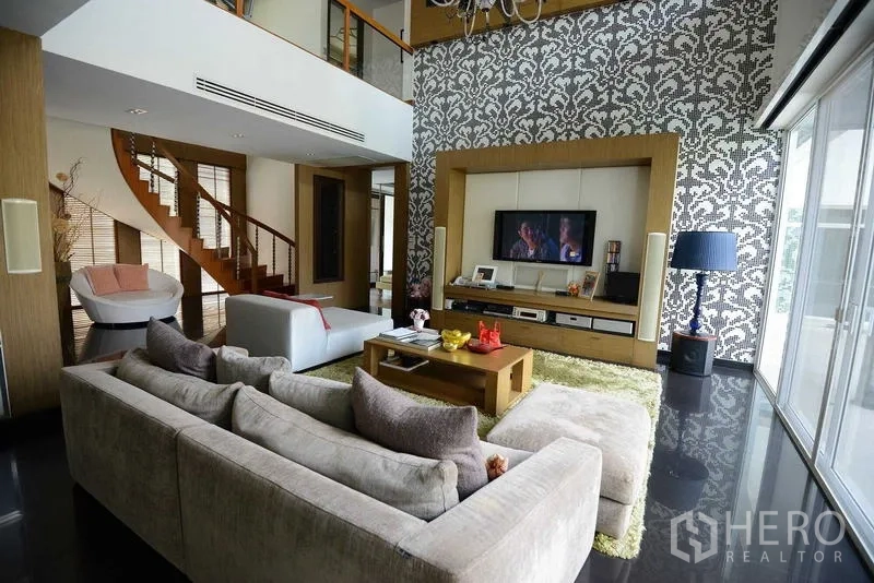 Detached House for sale in Bang Kapi, Bangkok - Double-height family room featuring patterned accent wall, TV unit and curved wooden staircase