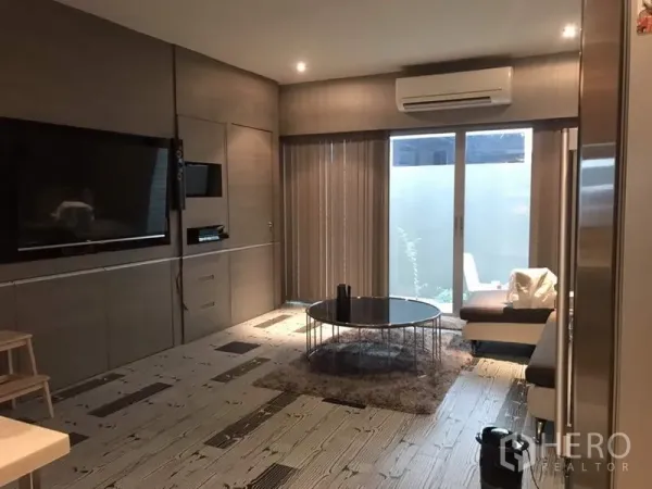 Townhouse for rent or sale in Suan Luang, Bangkok - Open-plan lounge with built-in TV wall, round glass coffee table, air conditioning, and vertical blinds over sliding glass doors.