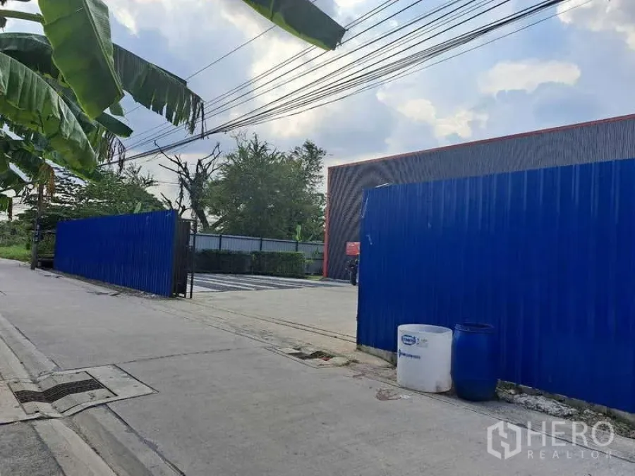 Warehouse for sale in Khlong Sam Wa, Bangkok - Street approach to the warehouse compound showing blue sliding gate, driveway entrance and overhead utilities.