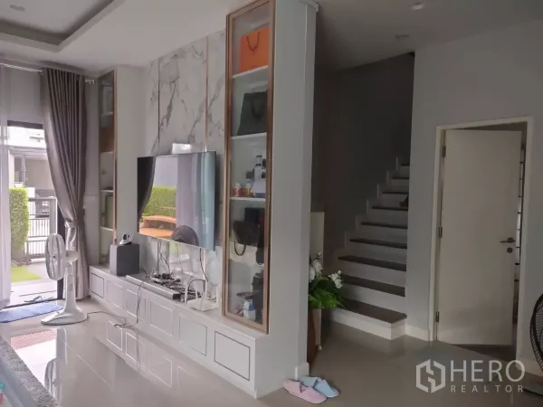 独立式住宅 出租或出售在Lam Luk Ka, Pathum Thani - 现代客厅，石纹电视墙、内置柜与楼梯，旁有推拉落地门。