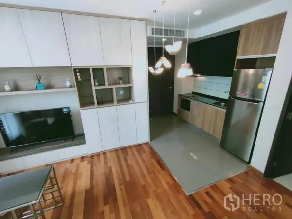 Condominium for rent in Ratchathewi, Bangkok - Modern living area with built-in cabinetry, pendant lights and a compact open kitchen with stainless fridge on warm wood floors.