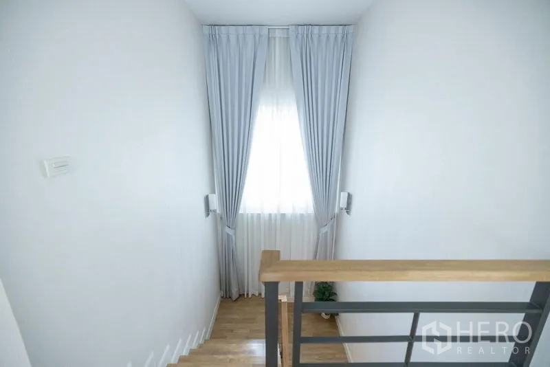 Detached House for sale in Prawet, Bangkok - Staircase landing with a tall window and light gray curtains allowing natural light.