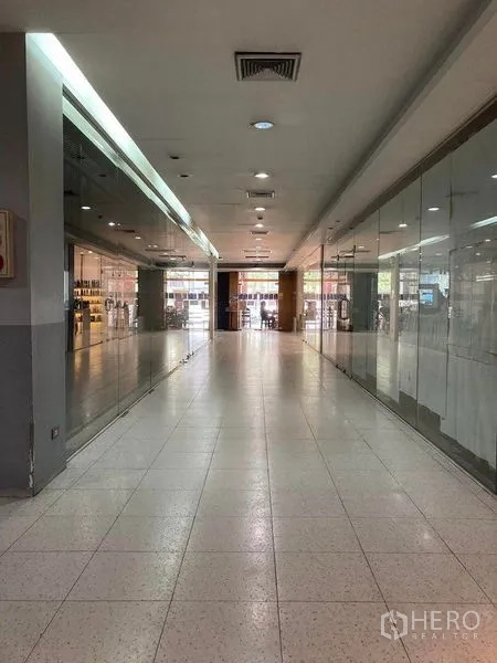 พื้นที่ขายปลีก ให้เช่า - ทางเดินห้างยาว มีหน้าร้านกระจกสองฝั่ง ไฟดาวน์ไลท์และช่องแอร์ มุ่งสู่ทางเข้า