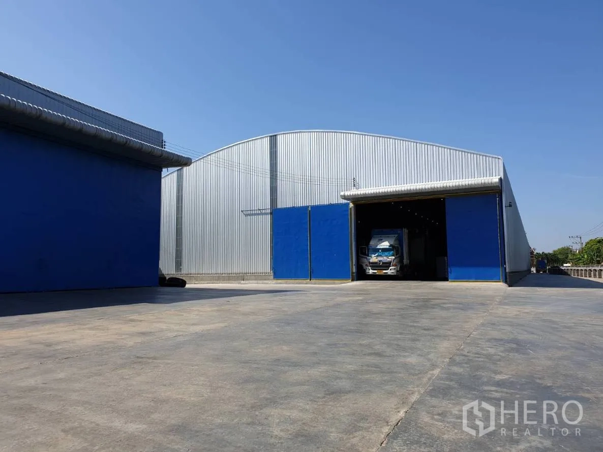 Warehouse for rent in Chaloem Phra Kiat, Saraburi - Steel-clad warehouse with blue sliding doors, high arched roof and wide concrete yard for truck access in Chaloem Phra Kiat, Saraburi.