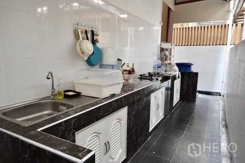 Detached House for sale in Bueng Kum, Bangkok - Practical Thai kitchen with long black countertop, sink, gas hob and white tiled walls.