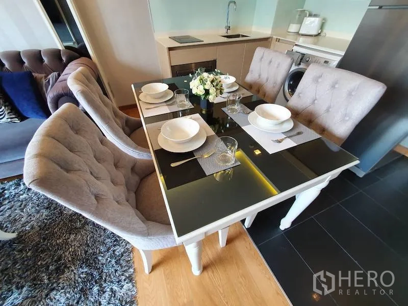 Condominium for rent in Watthana, Bangkok - Elegant four-seat dining table with tufted chairs beside a compact kitchen with cooktop and washing machine.