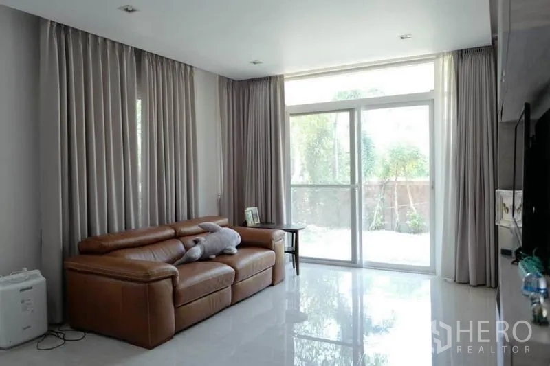 Detached House for sale in Pak Kret, Nonthaburi - Bright living room with large sliding glass doors, gray curtains and a brown leather sofa.