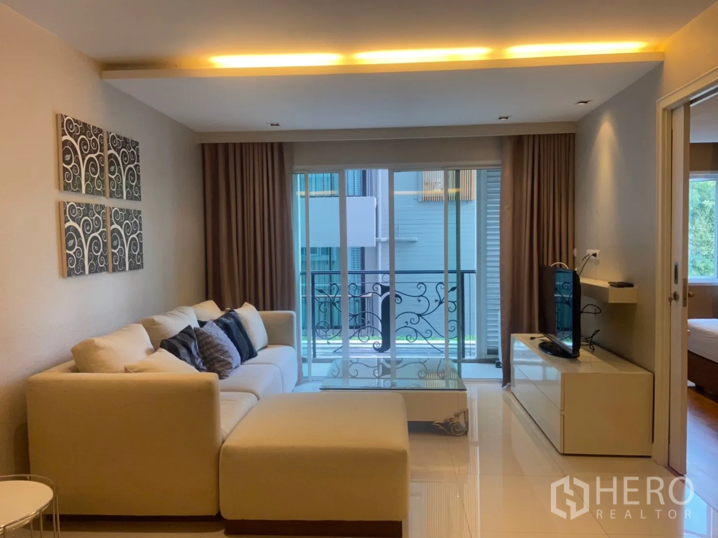 公寓 出租在Watthana, Bangkok - 客厅配L形沙发与电视柜，推拉门通向带铁艺栏杆的小阳台。