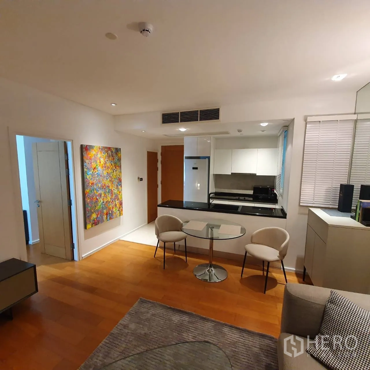 Condominium for rent in Watthana, Bangkok - Dining nook with round glass table beside a modern white kitchen and colorful wall art.