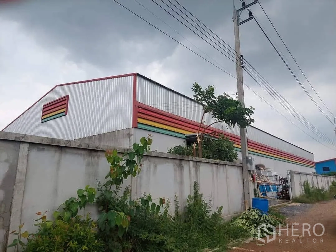 โกดัง for sale in Lam Luk Ka, Pathum Thani - มุมอาคารทรงจั่วสูง แผ่นผนังสีรุ้ง เสาไฟ และต้นไม้ริมรั้ว