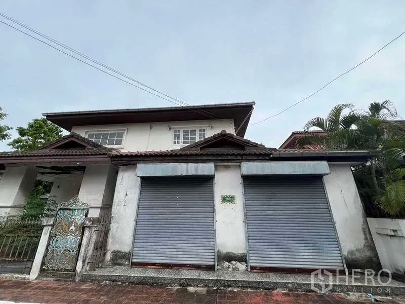 บ้าน ขายในPhra Khanong, Bangkok - ด้านหน้าบ้านใกล้ๆ แสดงประตูม้วนคู่ ชายคาหลังคากระเบื้อง และพืชทรอปิคัลข้างกำแพง