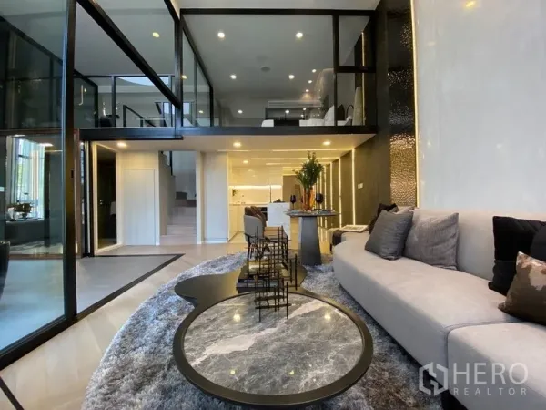 Detached House for sale in Watthana, Bangkok - Contemporary living room with sectional sofa, round coffee tables, and an open mezzanine over the kitchen.