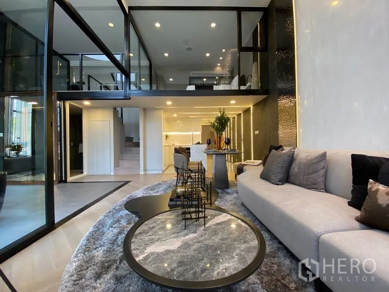 Detached House for sale in Watthana, Bangkok - Contemporary living room with sectional sofa, round coffee tables, and an open mezzanine over the kitchen.