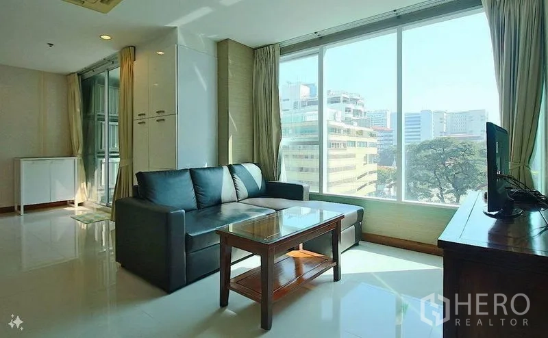 Condominium for sale in Watthana, Bangkok - Bright living room with large windows, city view, black sectional sofa and glass coffee table.