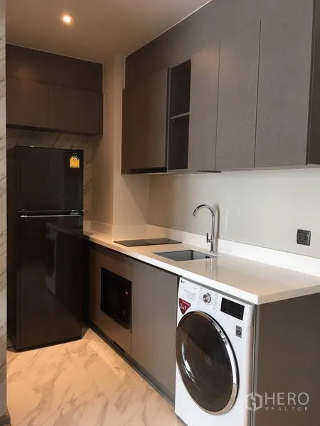 Condominium for rent in Watthana, Bangkok - Modern kitchen with dark cabinetry, refrigerator, cooktop and under-counter washer.