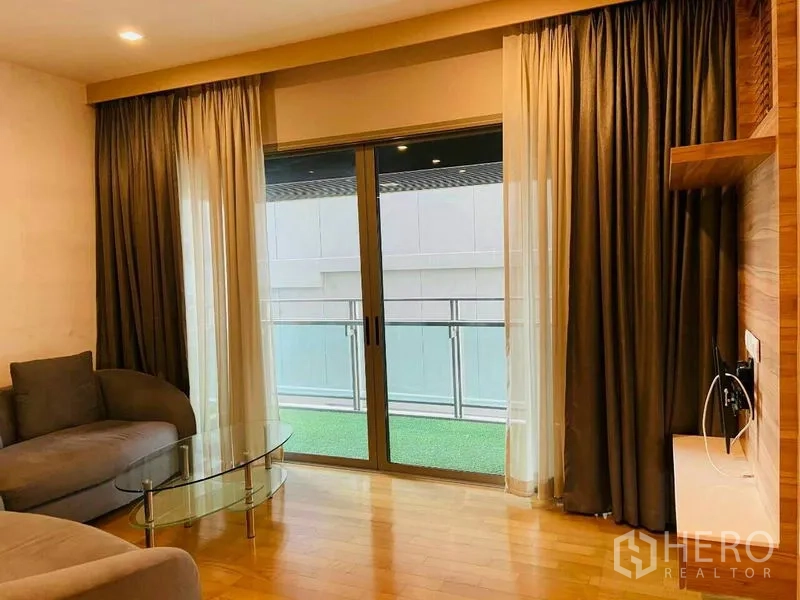Condominium for rent in Watthana, Bangkok - Cozy living room with sofa, glass coffee table, and sliding doors to a balcony with turf.