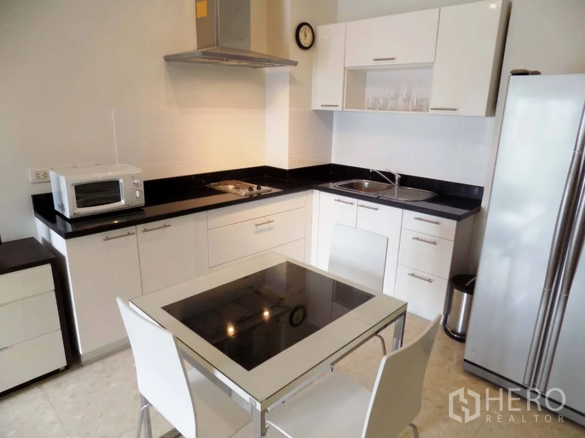 Condominium for rent or sale in Khlong Toei, Bangkok - Modern white kitchen with black countertops, glass-top dining table, cooktop, hood, microwave and tall fridge.