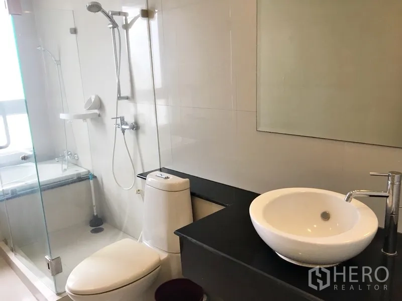 Condominium for rent in Khlong Toei, Bangkok - Bathroom angle showing vessel sink, countertop, toilet, and rainfall shower beyond glass.