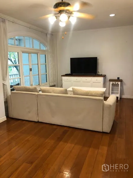 ทาวน์เฮาส์ ให้เช่าในMueang Samut Prakan, Samut Prakan - ห้องนั่งเล่นโซฟาตัวใหญ่หันเข้าทีวี ติดพัดลมเพดาน พื้นไม้ และประตูบานฝรั่งเศสออกสู่ระเบียง