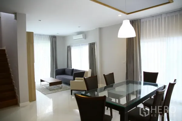 Detached House for rent or sale in Bang Na, Bangkok - Bright dining zone with glass table and six chairs beside a cozy lounge, staircase and full-height curtains.