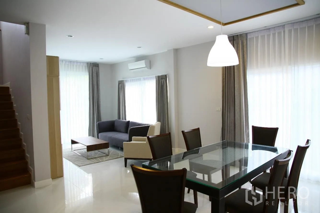 Detached House for rent or sale in Bang Na, Bangkok - Bright dining zone with glass table and six chairs beside a cozy lounge, staircase and full-height curtains.