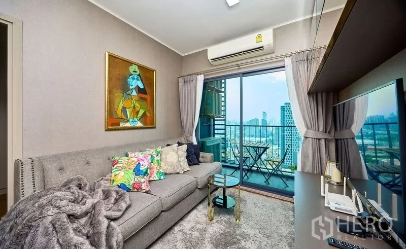 Condominium for rent in Phra Khanong, Bangkok - Cozy living room with gray sofa, wall art and sliding doors opening to a skyline-view balcony near BTS Bang Chak.