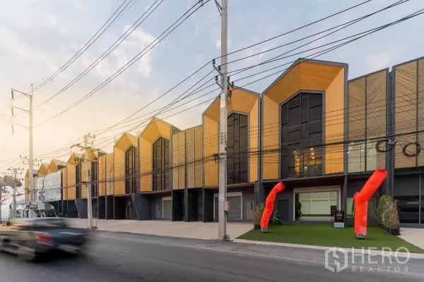 货栈 出租在Mueang Samut Sakhon, Samut Sakhon - 临街一排现代阁楼式仓库外立面，木质山墙与大玻璃，入口设红色气柱，傍晚光线柔和。