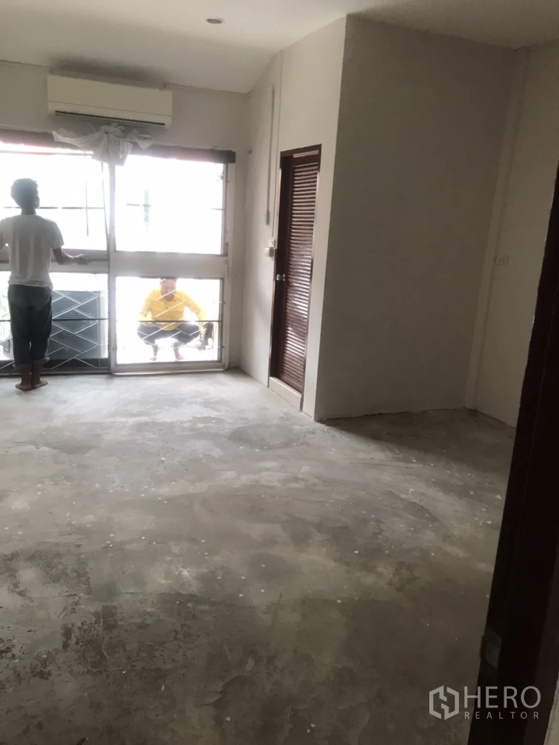 Home Office for rent in Khlong Toei, Bangkok - Bright room with air conditioner, large window with grille, unfinished floor, and a door to a side room.