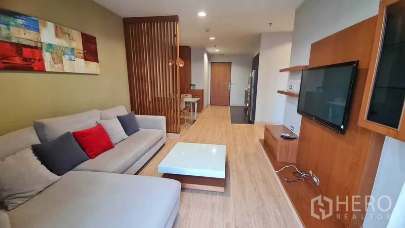 Condominium for rent or sale in Watthana, Bangkok - Living room with wall-mounted TV, shelving, sectional sofa, and partition leading to dining area.