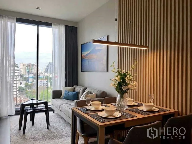 Condominium for rent in Watthana, Bangkok - Living area with sliding door to balcony, city view and a four‑seat dining table by a slatted wall.
